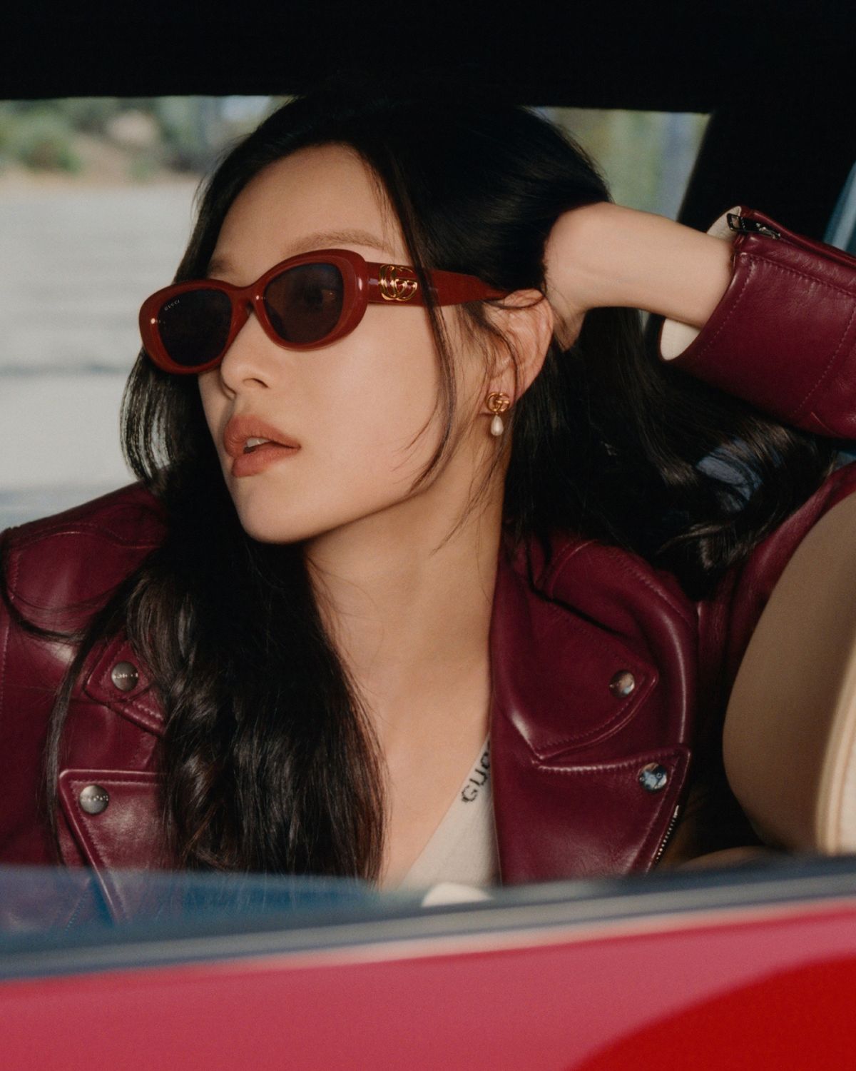 Femeie tânără cu păr lung negru purtând ochelari de soare Gucci cu ramă roșu-burgundy în stil cat-eye și detalii aurii, îmbrăcată într-o jachetă de piele burgundy și cercei cu perle, fotografiată în interiorul unei mașini pentru campania Gucci Eyewear Vară 2025